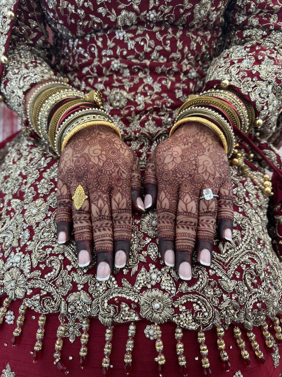 A bride's hennaed hands with stacked 22ct gold and glass bangles and a diamond ring, over a red and gold bridal outfit
